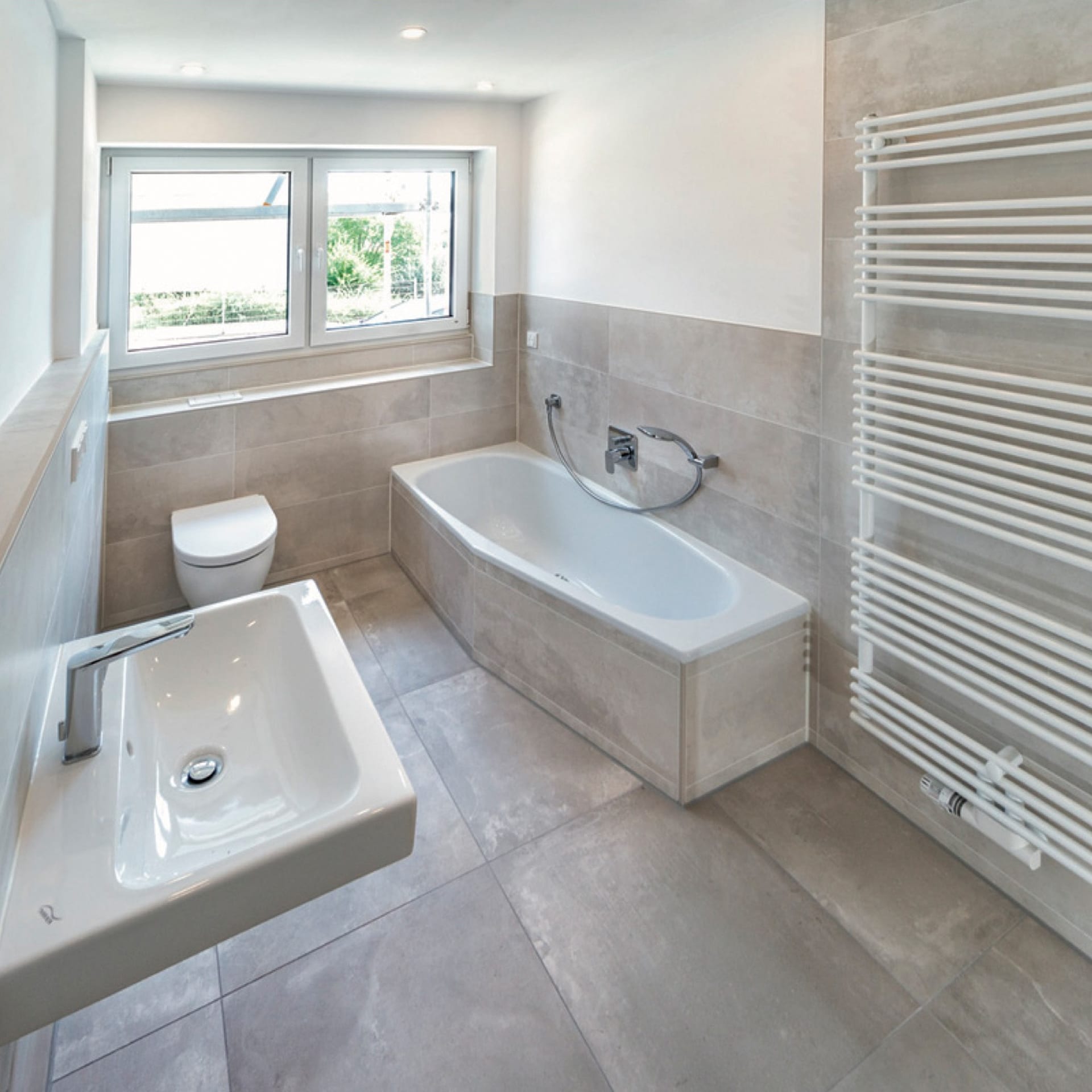 Modernes Badezimmer mit Fliesen, Badewanne, wandmontiertem Waschbecken, Toilette, beheiztem Handtuchhalter und großem Fenster